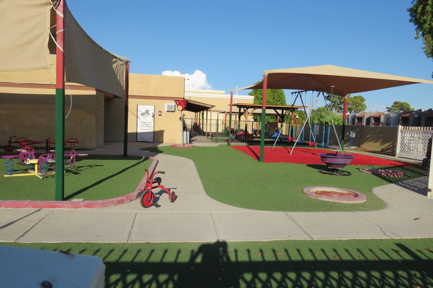 Nana&rsquo;s outdoor play yard: artificial turf, shade sails overhead,
 tricycles and play structures, empty between sessions.